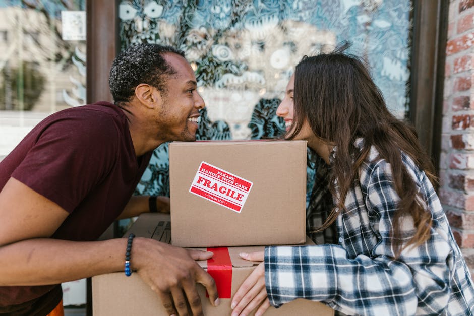 A man and a woman are outside near a brick building, engaged in a house removal process as part of a home relocation. The man is leaning forward with a smile, holding a cardboard box labeled 'Fragile' with red and white handling care stickers, indicating careful packing for furniture transport. The woman is also smiling and holding the same box, with several other packed cardboard boxes stacked in front of them, wrapped in red tape. The background shows a window with a decorative frosted glass design and a partially visible door, suggesting they are outside a residential property. The scene is well-lit with natural daylight, and the involved objects include cardboard boxes, packing tape, and the boxes themselves are made from brown kraft paper or cardboard, typical for a moving and packing process. This image relates to removals and moving services offered by [COMPANY_NAME], illustrating packing, handling fragile items, and the logistics involved in house removals, relevant to the context of avoiding hidden charges and ensuring smooth furniture transport.
