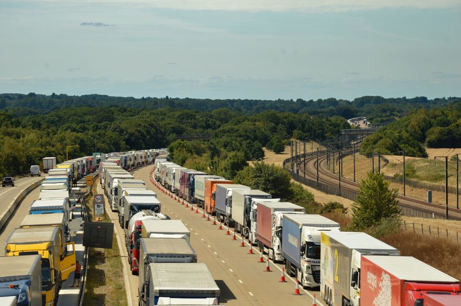 A line of parked cars and moving vehicles on a two-lane road during dusk, with some vehicles partially visible in the foreground and others further along the road, creating a congested traffic scene. 