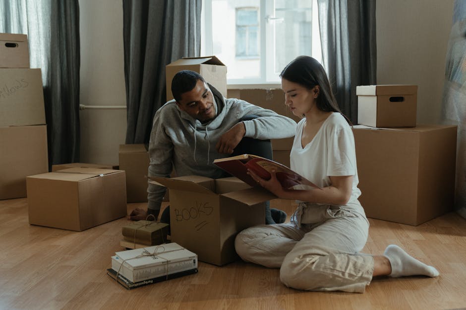 A young man and a young woman are inside a residential property, engaging in a home relocation process. The man, dressed in a grey hoodie, is kneeling on the floor beside a cardboard box labeled 'book