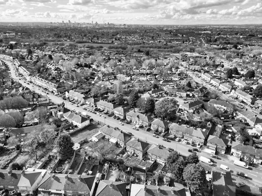 A black and white aerial photograph depicting a suburban residential area with a street lined with terraced and semi-detached houses, each with small gardens and driveways. Motor vehicles are parked a