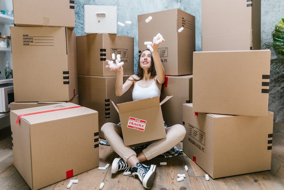 A woman sitting cross-legged on a wooden floor inside a home surrounded by numerous cardboard boxes, some sealed with red tape and others open. She is wearing a white tank top, beige pants, and black-and-white sneakers, and is smiling as she throws packing foam peanuts into the air. The boxes vary in size and have labels and markings, indicating their contents and destination within the house. The background includes a portion of a textured grey wall and a green potted plant, with natural indoor lighting illuminating the scene. This image showcases the packing process during a house relocation, with effective use of cardboard boxes, packing materials, and careful handling, reflecting the professional moving and removals services offered by Man and Van New Addington.