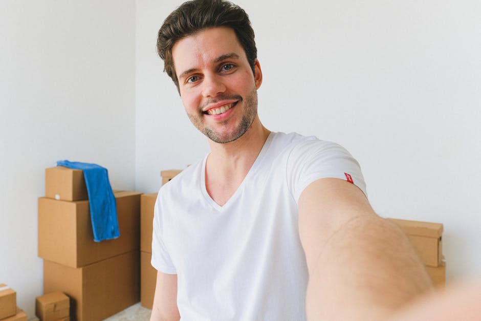 A young man with short brown hair and a friendly smile is taking a selfie inside a residential property, standing in front of a stack of cardboard moving boxes of various sizes. Some boxes are taped shut and a blue fabric cover is draped over one of them. The man is wearing a white T-shirt and appears to be engaged in a home relocation process, potentially part of a furniture transport or packing and moving service. Behind him, the interior features a plain white wall and a light-colored overall environment, indicating a clean and organized space prepared for a house move. The lighting is bright, emphasizing the details of the boxes and the man's face, reflecting a professional yet casual atmosphere typical of residential removals carried out by companies like Man and Van New Addington during the loading or packing stage.