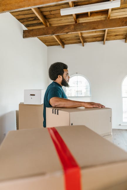A woman sitting cross-legged on a wooden floor inside a home surrounded by numerous cardboard boxes, some sealed with red tape and others open. She is wearing a white tank top, beige pants, and black-and-white sneakers, and is smiling as she throws packing foam peanuts into the air. The boxes vary in size and have labels and markings, indicating their contents and destination within the house. The background includes a portion of a textured grey wall and a green potted plant, with natural indoor lighting illuminating the scene. This image showcases the packing process during a house relocation, with effective use of cardboard boxes, packing materials, and careful handling, reflecting the professional moving and removals services offered by Man and Van New Addington.