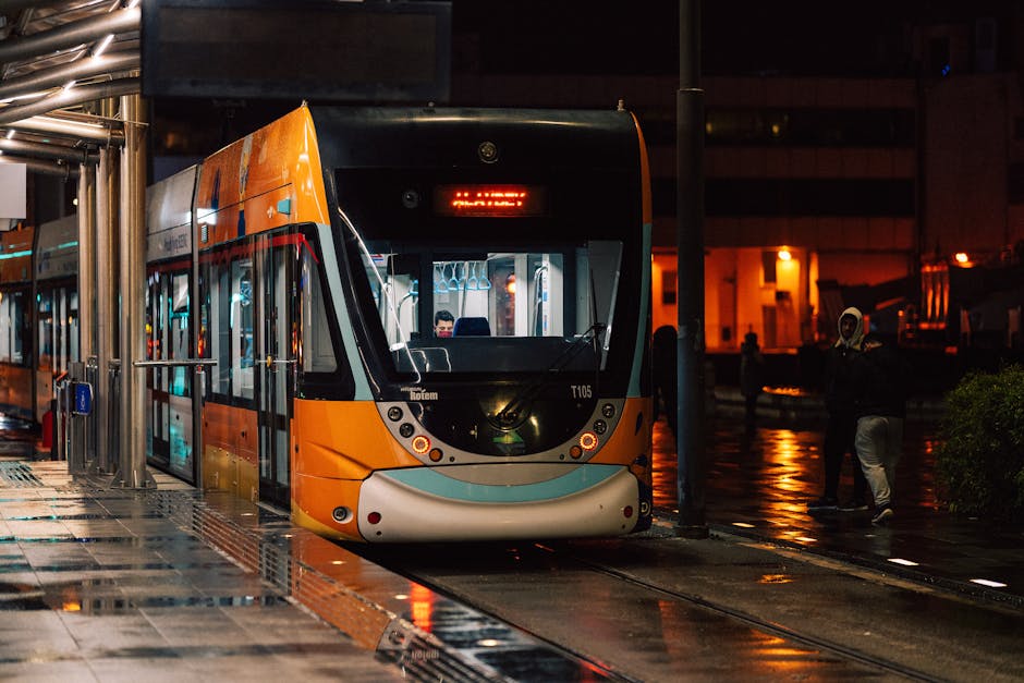 A modern orange and black tram is stationary at a tram stop during nighttime, with its front facing the camera and illuminated interior lights visible through the large front window. The tram is positioned on tracks embedded in a wet pavement reflecting nearby streetlights and surrounding buildings. To the left, a sheltered area with metal support beams and a glass enclosure is visible, possibly for passenger waiting. Several pedestrians are seen nearby, some wearing hooded jackets, indicating cool or rainy weather. In the background, an urban environment with illuminated windows and street lighting creates a warm glow against the dark sky. The scene captures a typical city transport setting, where Man and Van New Addington may coordinate home relocation or furniture transport services, especially considering the context of moving logistics associated with house removals and packing activities.