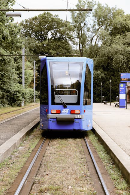 A modern blue tram parked on narrow tracks at a tram station, viewed from the rear with large tinted windows reflecting surrounding trees and sky. The tram has a white and blue logo on the back, with a small number plate displaying '512'. Overhead, there are electrical wires supported by metal poles, extending across the rail line. To the right of the tram, there are blue and white ticket machines and a seating area with benches, set on a paved platform. The station is surrounded by greenery, including tall trees and bushes, creating a semi-enclosed environment. The overall scene suggests a quiet urban or suburban tram stop, with no visible passengers, and the setting appears suitable for a home relocation or furniture transport process, as part of a professional removal service by Man and Van New Addington.