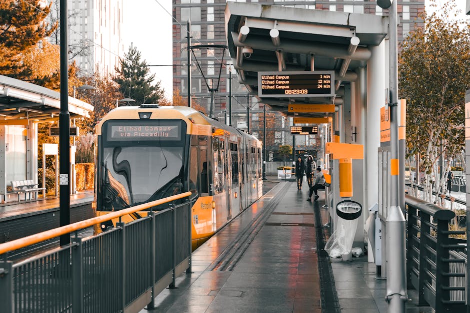 A modern orange and black tram is stationary at a tram stop during nighttime, with its front facing the camera and illuminated interior lights visible through the large front window. The tram is positioned on tracks embedded in a wet pavement reflecting nearby streetlights and surrounding buildings. To the left, a sheltered area with metal support beams and a glass enclosure is visible, possibly for passenger waiting. Several pedestrians are seen nearby, some wearing hooded jackets, indicating cool or rainy weather. In the background, an urban environment with illuminated windows and street lighting creates a warm glow against the dark sky. The scene captures a typical city transport setting, where Man and Van New Addington may coordinate home relocation or furniture transport services, especially considering the context of moving logistics associated with house removals and packing activities.