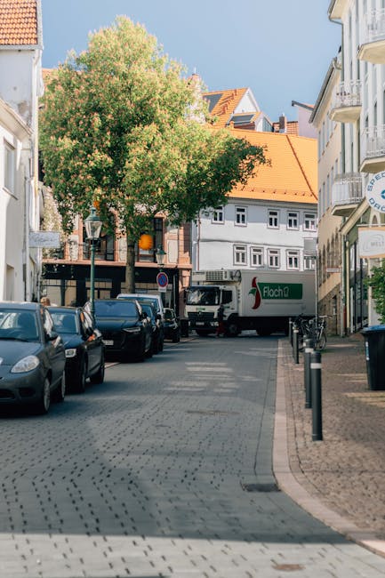 A narrow urban street scene with cobblestone paving, lined by multi-storey white and beige residential and commercial buildings with balconies. A large leafy tree with green foliage is centered, partially shading the area. Parked cars are along both sides of the street, including a silver vehicle directly in front of a small man and van service truck with a green logo, positioned near the curb. The van is being loaded or unloaded, with the rear doors open, indicating a home relocation or furniture transport process. Equipment such as black bollards prevent parking on the sidewalk, and a bicycle is secured to a stand nearby. The scene is well-lit, with sunlight casting shadows, and the overall setting depicts a busy, residential area suitable for house removals and moving logistics, as handled by companies like Man and Van New Addington.