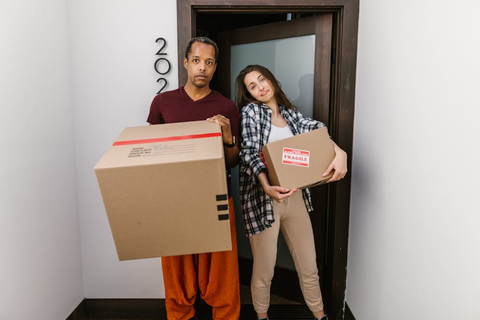 A man and woman standing inside a doorway, each holding a cardboard box for home relocation. The man, dressed in a maroon t-shirt and orange pants, is carrying a large box with black tape and several labels visible on its surface. The woman, wearing a checkered shirt over a white top and beige trousers, is holding a smaller box labeled 