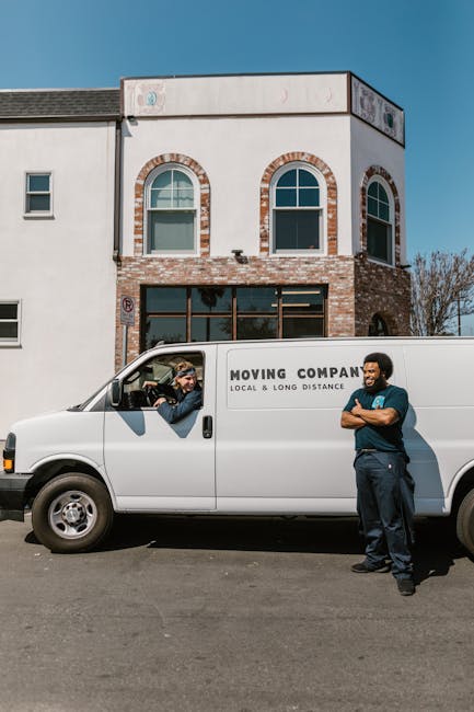 A white moving van parked in front of a two-storey residential building with a brick and white exterior under a clear blue sky. The van has the words 'Moving Company, Local & Long Distance' printed on the side. Inside the driver’s cabin, a person wearing sunglasses and a cap is visible, engaged in conversation with a second person standing outside the van. The second individual is dressed in dark work clothes, with arms crossed, and appears to be preparing for a home relocation or furniture transport. The building features arched windows on the upper floor and large glass doors on the ground floor, with some cardboard boxes and packing materials visible through the windows, indicating recent packing or moving preparations. The scene is set on a paved street, and the overall context suggests a professional moving process involving careful loading or unloading near the property, consistent with house removals and relocation services.