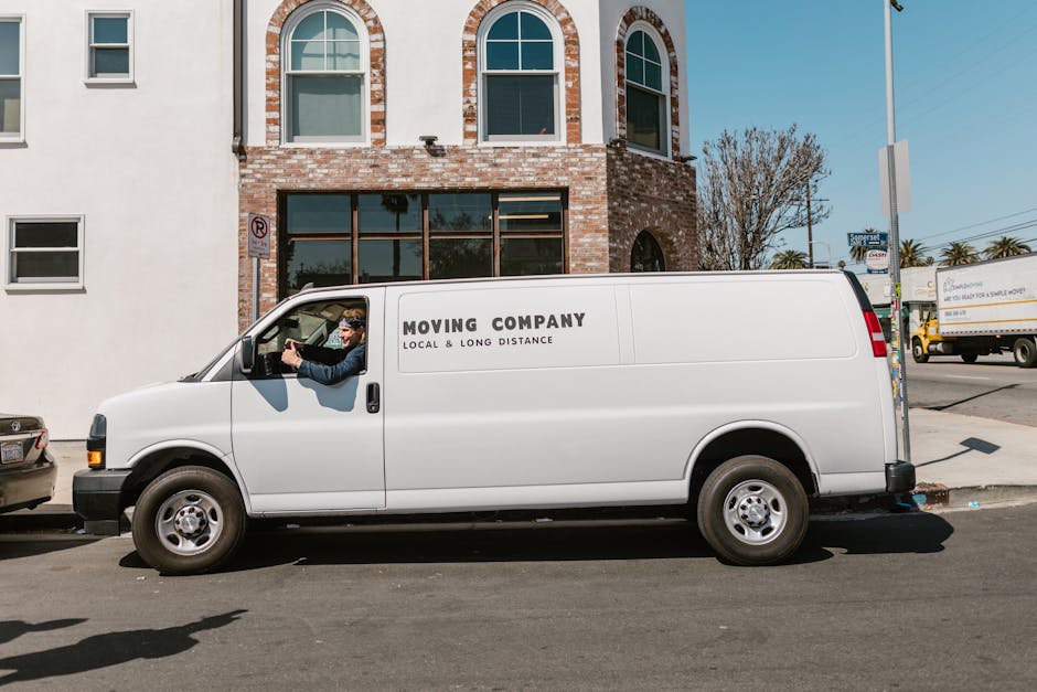A white moving van parked in front of a two-storey residential building with a brick and white exterior under a clear blue sky. The van has the words 'Moving Company, Local & Long Distance' printed on the side. Inside the driver’s cabin, a person wearing sunglasses and a cap is visible, engaged in conversation with a second person standing outside the van. The second individual is dressed in dark work clothes, with arms crossed, and appears to be preparing for a home relocation or furniture transport. The building features arched windows on the upper floor and large glass doors on the ground floor, with some cardboard boxes and packing materials visible through the windows, indicating recent packing or moving preparations. The scene is set on a paved street, and the overall context suggests a professional moving process involving careful loading or unloading near the property, consistent with house removals and relocation services.
