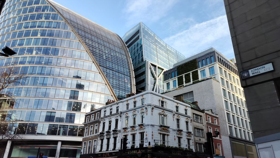 A city street scene showing a combination of architectural styles, with modern glass office buildings towering above an older, white-painted brick building with decorative moldings and multiple windows. The modern buildings feature reflective glass facades, curved and angular designs, and some greenery on their rooftops. The older building at street level has a sign reading 'LONDON WALL' on a gray concrete wall on the right side. The sky is partly cloudy with blue patches visible, and natural daylight illuminates the scene. The image captures an urban environment that could be associated with house removals, property differences, and moving logistics within a city context, relevant to relocation services offered by companies like Man and Van New Addington.
