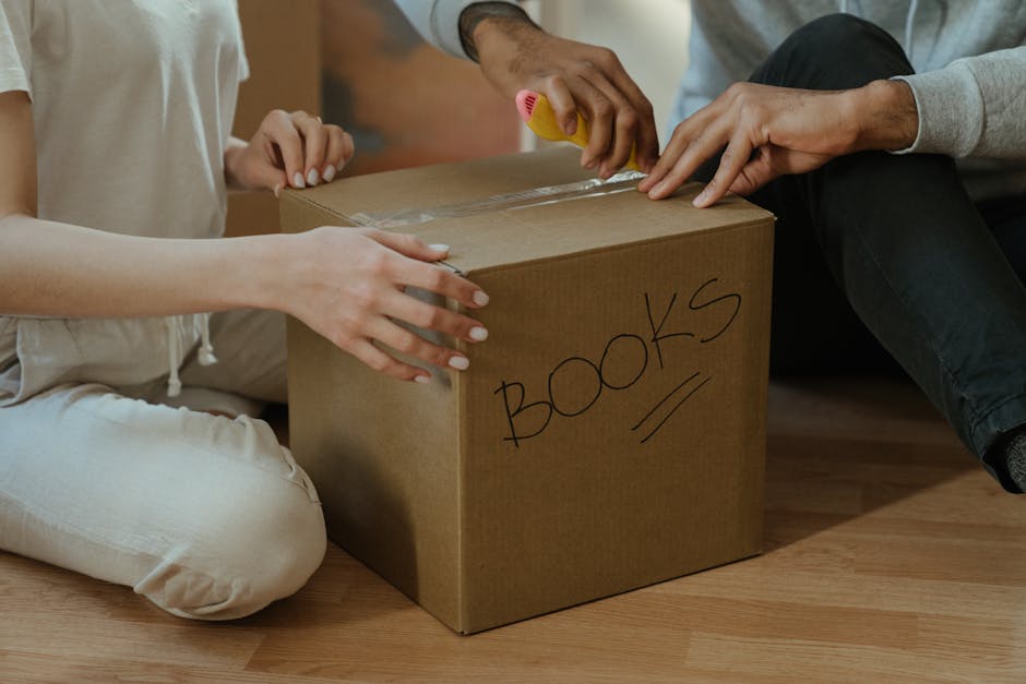 Two individuals are sitting on a wooden floor inside a house, preparing to transport a cardboard box labeled 'BOOKS' in black marker. One person is holding the box steady with their left hand, while the other is using a yellow marker to write or label it. The box is sealed with clear packing tape along the top edges. The scene captures the packing process during a home relocation, with the focus on organizing and boxing up belongings. In the background, part of a doorway and the interior wall are visible, indicating the inside of a residential property. The individuals are dressed casually, and the lighting appears natural, suggesting daylight. This image reflects the packing stage of furniture transport and moving logistics, emphasizing careful handling and organization during a house move, fitting for a professional removals service like Man and Van New Addington.