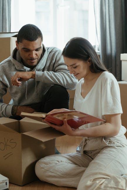 Two individuals, a man and a woman, are inside a residential property preparing for a home relocation. The woman is sitting on the floor, holding a red blanket or packaging material, while the man, sitting beside her, looks on and is engaged in the packing process. Surrounding them are cardboard boxes, one of which is labeled with handwritten text, likely indicating contents for moving. The background includes a window with light curtains, allowing natural daylight to illuminate the scene, and a window sill with some objects. The scene captures the packing stage of furniture transport and moving logistics, emphasizing careful handling of household items before loading them onto a van, which is part of the house removals process handled by [COMPANY_NAME]. This detailed description reflects the meticulous planning involved in professional removals, ensuring safe packing and transport in home relocation projects.