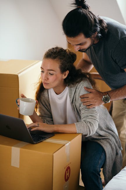 Two individuals are sitting on a wooden floor inside a house, preparing to transport a cardboard box labeled 'BOOKS' in black marker. One person is holding the box steady with their left hand, while the other is using a yellow marker to write or label it. The box is sealed with clear packing tape along the top edges. The scene captures the packing process during a home relocation, with the focus on organizing and boxing up belongings. In the background, part of a doorway and the interior wall are visible, indicating the inside of a residential property. The individuals are dressed casually, and the lighting appears natural, suggesting daylight. This image reflects the packing stage of furniture transport and moving logistics, emphasizing careful handling and organization during a house move, fitting for a professional removals service like Man and Van New Addington.