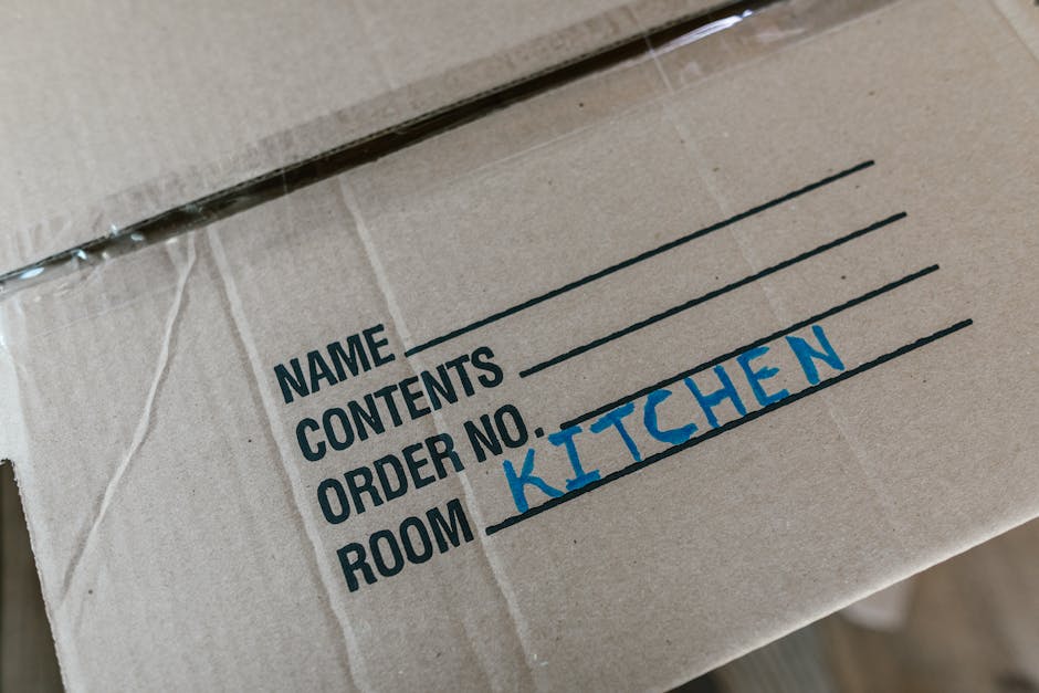 Close-up of a cardboard moving box labelled with black printed text and blue handwritten text indicating it contains kitchen items, with the word 'KITCHEN' prominently written. The box is sealed with clear packing tape and is situated in a home interior, likely on a staircase or hallway, ready for packing or transport during a home relocation. The environment suggests an interior space involved in furniture transport and packing for a house move, with other boxes or packing materials possibly out of view. This scene illustrates the process of packing and moving, supported by the services of Man and Van New Addington as part of their removals assistance within the context of house relocation.