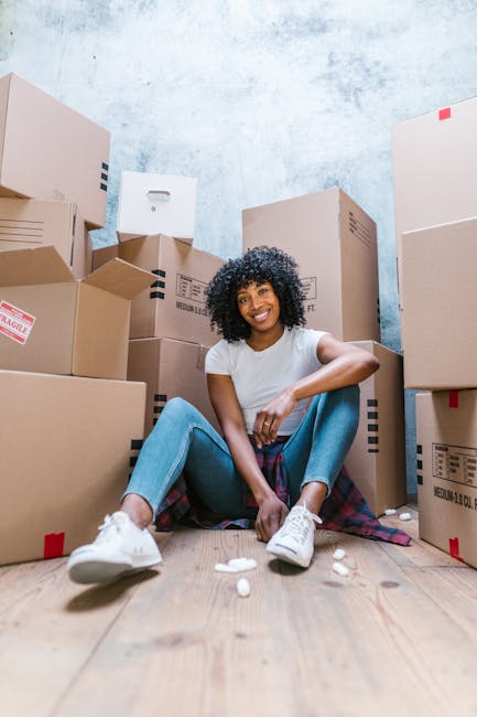 A young woman with curly black hair and a bright smile is seated on a wooden floor surrounded by several cardboard boxes of various sizes, some sealed with red or black tape and others open. She is wearing a white t-shirt, light blue jeans, and white sneakers, with a plaid shirt tied around her waist. The boxes contain household items, and a few foam packing peanuts are scattered on the floor nearby. The background features a plain, light blue wall with a slightly textured surface, indicating an indoor space ready for household packing and home relocation. This scene reflects the packing stage of a house move, with the woman in a relaxed pose amid her packed belongings, illustrating the work involved in furniture transport and moving logistics, an area serviced by Man and Van New Addington.