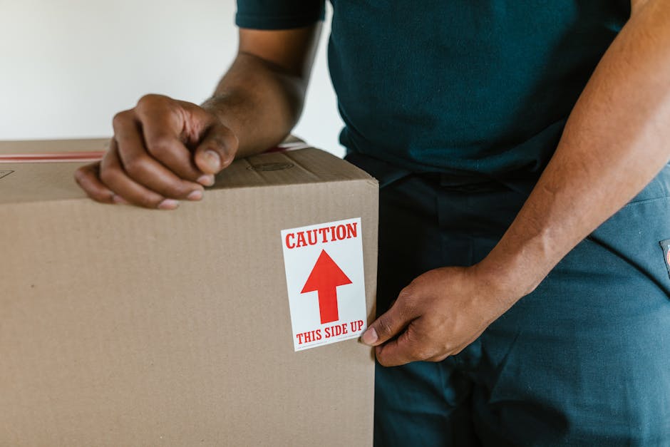 Close-up of a cardboard moving box labelled with black printed text and blue handwritten text indicating it contains kitchen items, with the word 'KITCHEN' prominently written. The box is sealed with clear packing tape and is situated in a home interior, likely on a staircase or hallway, ready for packing or transport during a home relocation. The environment suggests an interior space involved in furniture transport and packing for a house move, with other boxes or packing materials possibly out of view. This scene illustrates the process of packing and moving, supported by the services of Man and Van New Addington as part of their removals assistance within the context of house relocation.