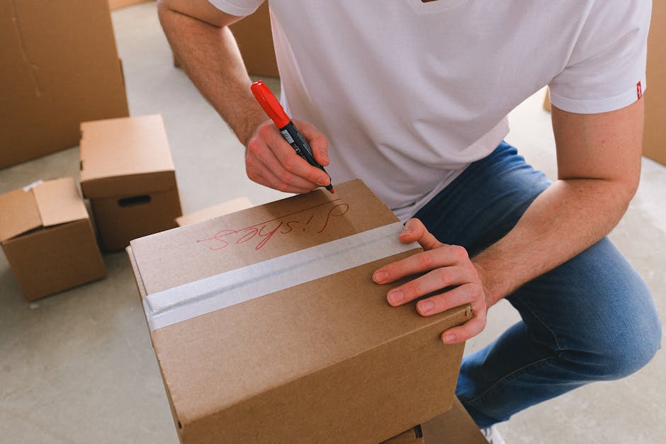 A person wearing a white t-shirt and blue jeans is kneeling on a light-colored floor inside a home or storage area, using a red marker to write the word 'Tips' on a medium-sized cardboard moving box. The individual is grasping the box firmly with their left hand while writing with their right hand. Several other cardboard boxes of various sizes, some closed and some open, are stacked or placed nearby, indicating packing for a home relocation. The boxes are made of brown corrugated cardboard, with some sealed with white packing tape. The setting appears well-lit, likely with natural or artificial light illuminating the workspace, and the scene depicts a typical packing or moving process managed by specialist removals services such as Man and Van New Addington, as part of furniture transport and packing arrangements during a house move.