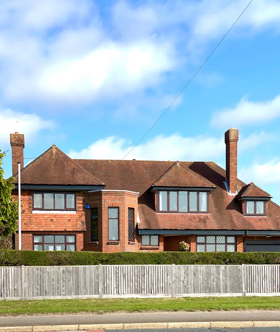 A large, multi-story brick house with a sloped red-tile roof is visible behind a wooden fence, situated on a suburban street. The house features multiple large rectangular windows with dark frames, some with decorative brickwork around them, and a tall brick chimney on the right side. The front yard has well-maintained green lawns with a neatly trimmed hedge along the base of the house. A blue planet-shaped ornament is mounted near the centre window on the left side of the house. The sky above is partly cloudy with patches of blue, indicating daytime weather. The scene is captured during a house removal or relocation process, as furniture and packing materials are not visible in the image, but the house's appearance suggests a home suitable for professional removals services by companies like Man and Van New Addington, who specialise in house removals and transport of bulk furniture during home relocations.