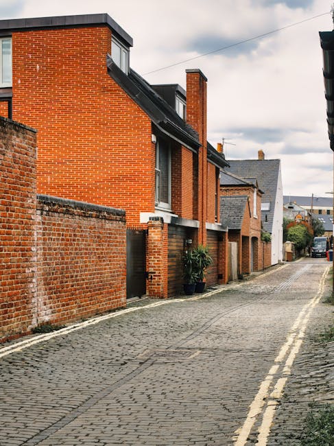 A narrow residential street with cobblestone pavement, lined with red-brick terraced houses featuring small front gardens and black doorways. Some houses have potted plants near entrance points. The street is relatively quiet with no visible pedestrians, and parked vehicles are seen further down the road. The sky is overcast, suggesting cloudy weather. This location appears suitable for house removals and furniture transport, with clear access for moving services such as the ones provided by Man and Van New Addington, who typically handle packing, loading, and home relocation logistics on such narrow lane jobs.